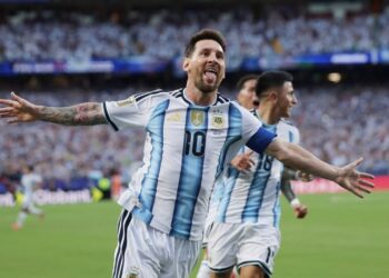 Argentine captain Lionel Messi, with his arms outstretched and sticking out his tongue in a spirited celebration, wearing the traditional white and light blue No. 10 shirt, after scoring for his country. A blurry teammate (number 16) is behind him, against a backdrop of a packed stadium crowd.