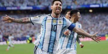 Argentine captain Lionel Messi, with his arms outstretched and sticking out his tongue in a spirited celebration, wearing the traditional white and light blue No. 10 shirt, after scoring for his country. A blurry teammate (number 16) is behind him, against a backdrop of a packed stadium crowd.