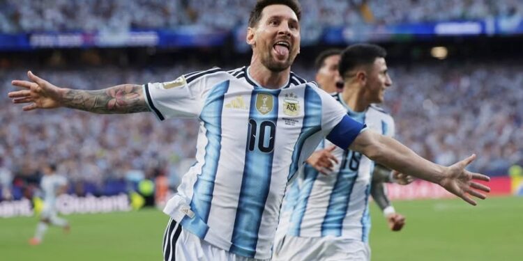 Argentine captain Lionel Messi, with his arms outstretched and sticking out his tongue in a spirited celebration, wearing the traditional white and light blue No. 10 shirt, after scoring for his country. A blurry teammate (number 16) is behind him, against a backdrop of a packed stadium crowd.