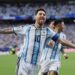 Argentine captain Lionel Messi, with his arms outstretched and sticking out his tongue in a spirited celebration, wearing the traditional white and light blue No. 10 shirt, after scoring for his country. A blurry teammate (number 16) is behind him, against a backdrop of a packed stadium crowd.